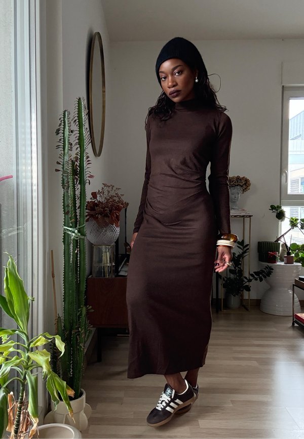 Abito lungo marrone aderente con maniche lunghe, realizzato in tessuto morbido. Abbinato a sneakers scure e accessori. Ambientazione interna con piante.