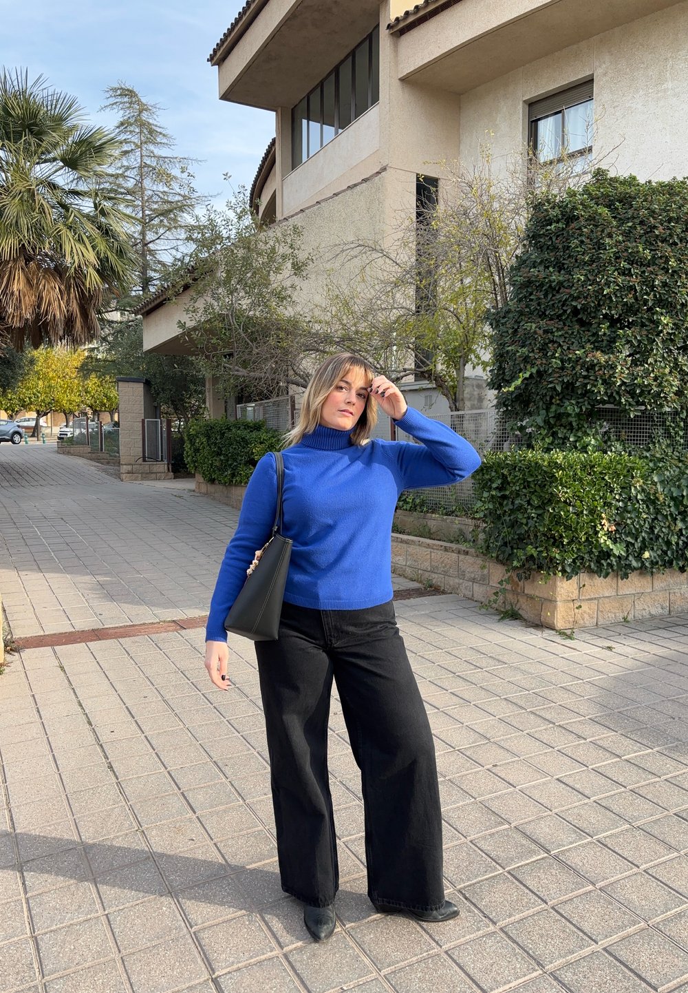 Femme en col roulé bleu et pantalon noir debout sur une rue pavée avec de la verdure et un bâtiment beige en arrière-plan, tenant un sac à main noir.