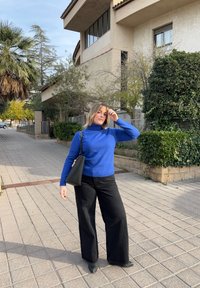 Femme en col roulé bleu et pantalon noir debout sur une rue pavée avec de la verdure et un bâtiment beige en arrière-plan, tenant un sac à main noir.
