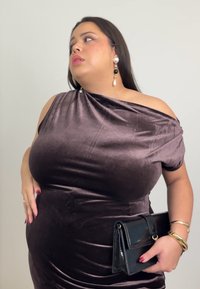 Samta brūna kleita ar plecu atsegumu, ar savilkumu detaļām un sp�īdīgu melnu rokassomu. Modelis valkā auskarus ar pērļu akcentiem un aproci.