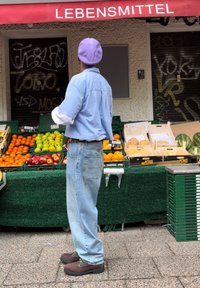Une personne portant une chemise rayée bleu clair et un bonnet violet se tient près d'un stand de fruits avec des caisses vertes, exposant des fruits colorés comme des oranges et des pommes.