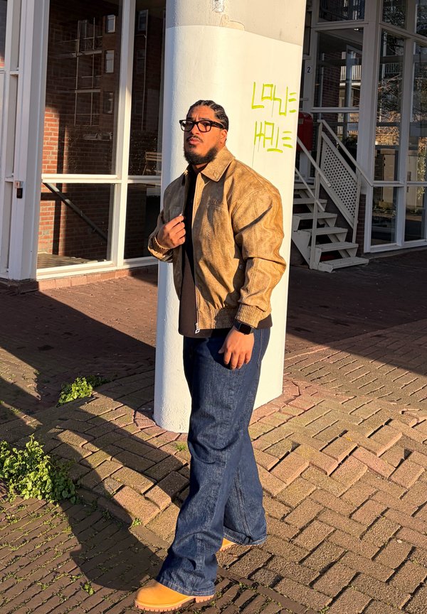 Mann med flettehår og briller har på seg en beige jakke, mørke jeans og støvler, står nær en hvit søyle med gul graffiti utenfor en bygning.