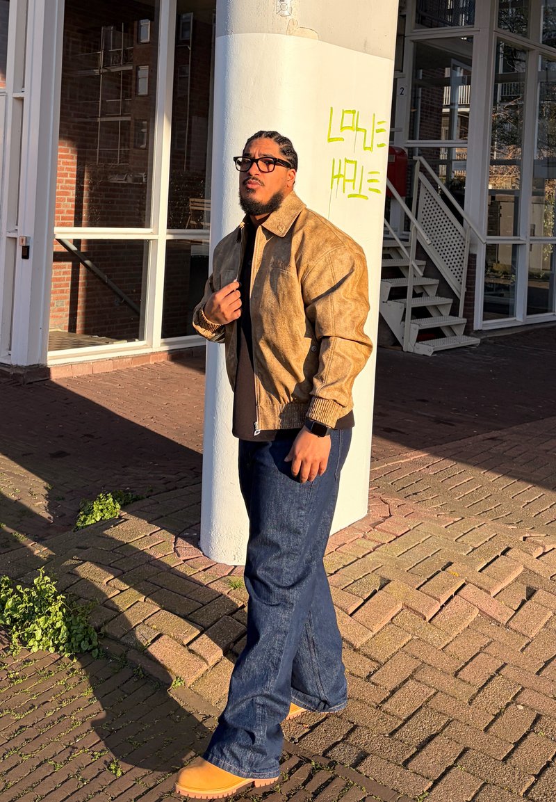 Homme aux cheveux tressés et lunettes porte une veste beige, un jean foncé et des bottes, debout près d'un pilier blanc avec des graffitis jaunes à l'extérieur d'un bâtiment.