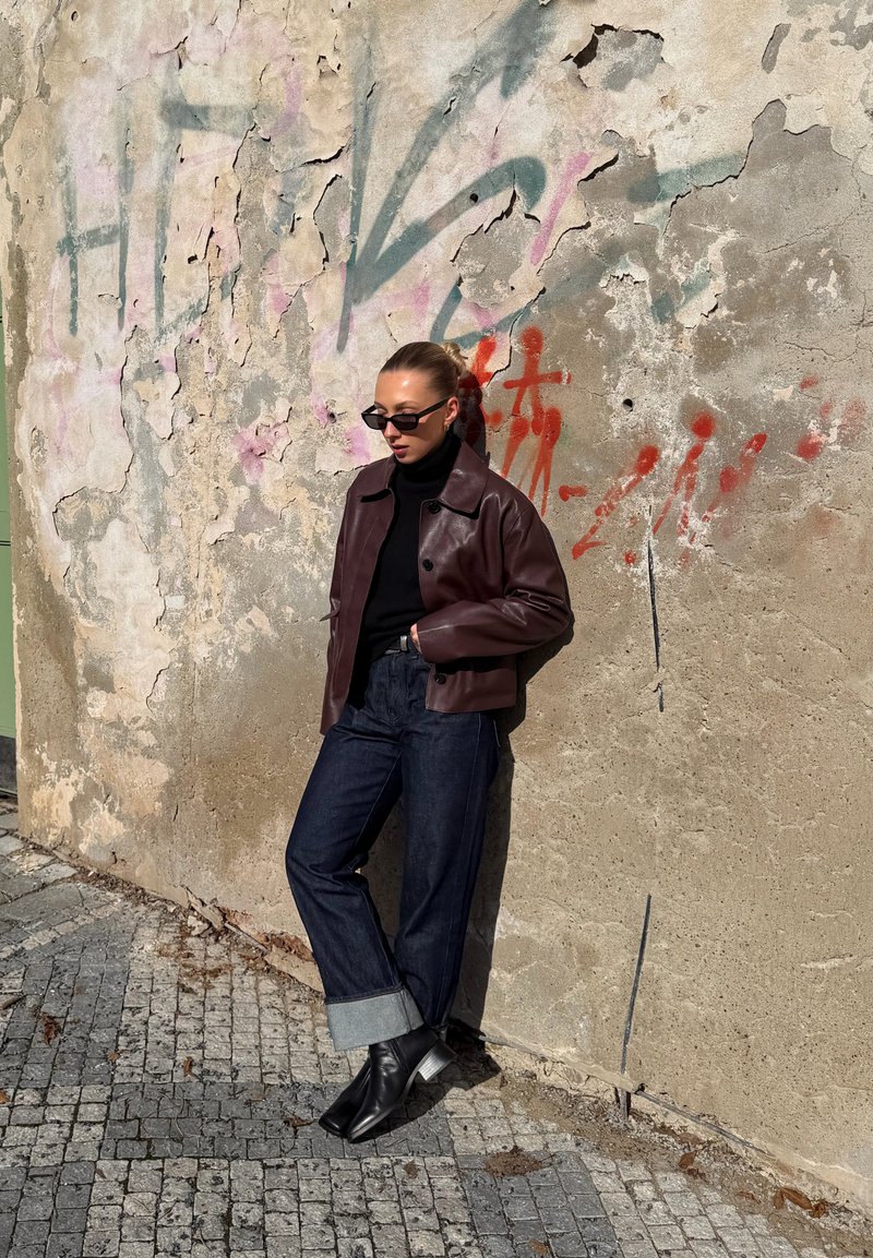 Femme en col roulé noir, veste en cuir bordeaux, jean sombre retroussé et bottes, appuyée contre un mur usé couvert de graffiti au soleil.