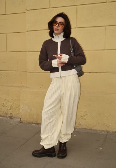Suéter de punto marrón y blanco con cremallera, pantalones de pierna ancha de rayas crema y botas de tobillo marrones, con grandes gafas de sol naranjas y un bolso pequeño.