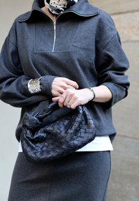 Femme tenant un sac à main en cuir tressé noir, portant un pull zippé gris foncé, une jupe assortie, un bracelet manchette argenté et des bagues.