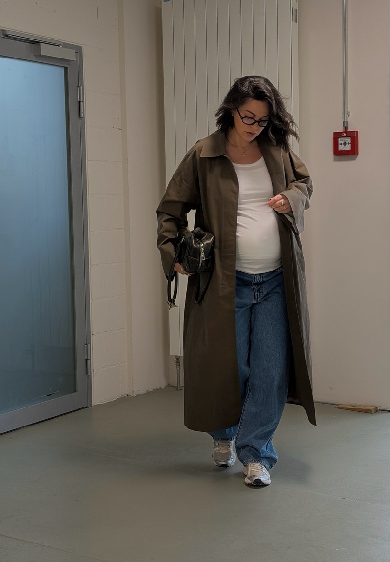 Femme enceinte portant des lunettes, un long manteau marron, un haut blanc et un jean, marchant à l'intérieur en tenant un sac à main noir.