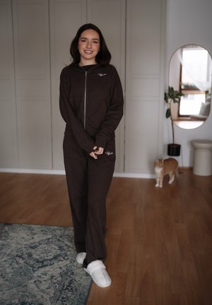 Joven sonriente, vestida con un chándal marrón oscuro y zapatillas blancas, de pie sobre el suelo de madera cerca de una alfombra y un gato naranja junto a un espejo.