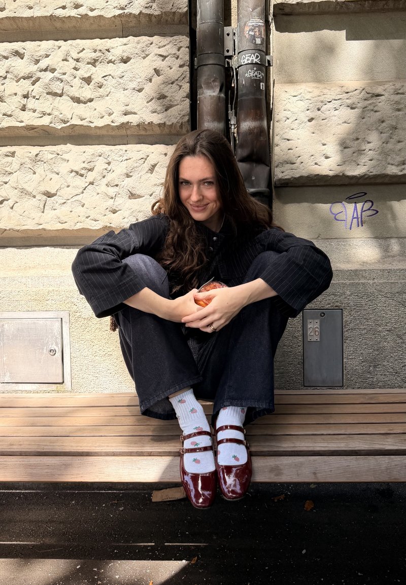 Jeune femme aux longs cheveux bruns, portant une veste sombre à fines rayures et un pantalon noir, assise jambes croisées sur un banc contre un mur en pierre.
