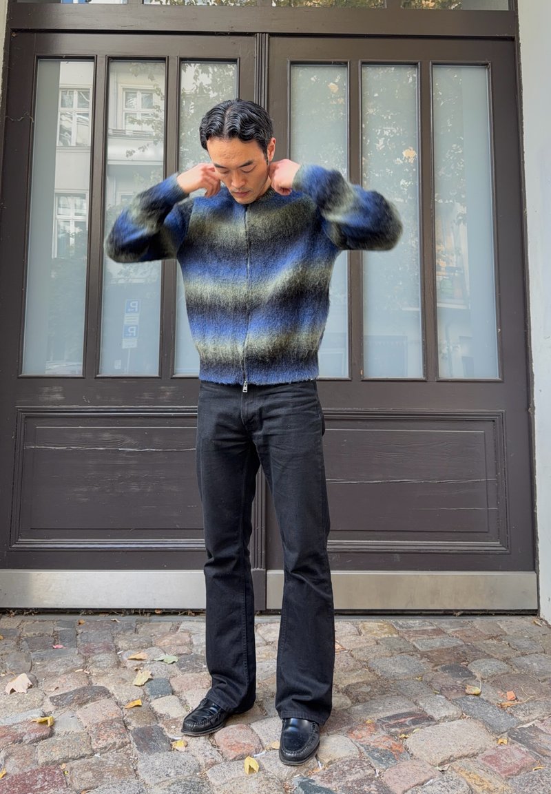 Homme ajustant le col d’un pull zippé à rayures bleues et vertes, debout sur des pavés devant des portes doubles sombres avec des panneaux en verre.