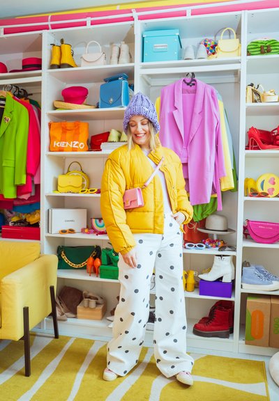 Mujer con chaqueta amarilla, gorro de lana morado y pantalones blancos con lunares, frente a un armario abierto con ropa colorida, zapatos y bolsos.