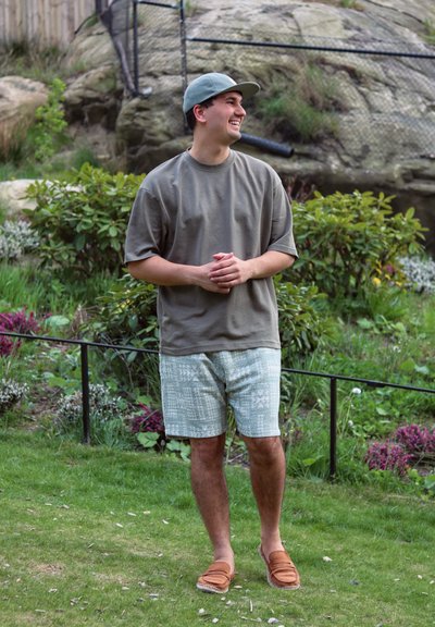 Camiseta de manga corta color verde oliva, pantalones cortos de color verde claro con estampado y zapatos marrones tipo slip-on, situados en un área exterior con césped y plantas.