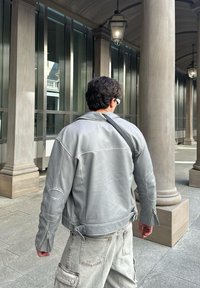 Γκρι δερμάτινο μπουφάν με διαρθρωτικούς ώμους και φερμουάρ στους καρπούς, συνδυασμένο με ανοιχτό γκρι παντελόνι cargo. Το σκηνικό διαθέτει πέτρινες κολώνες και γυαλί.