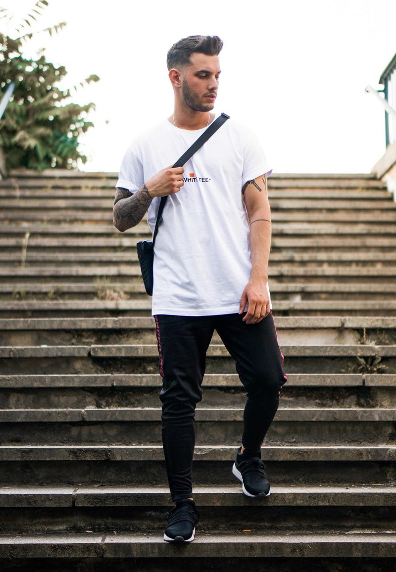 Weißes T-Shirt mit dem Text "WHITEEE", schwarze Jogginghose mit kastanienbraunen Seitenstreifen, schwarze Sneakers, die steinige Treppen heruntergehen.