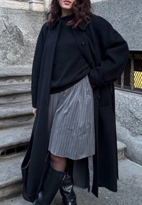 Cappotto lungo nero sopra un maglione nero, abbinato a una gonna grigia a righe e stivali alti neri fino al ginocchio. Tessuto texture e vestibilità comoda.