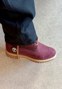 Bordeaux ruskinds ankelstøvler med gummisål, cirkulært logo-patch og lysebrune snørebånd, placeret på en beige betonoverflade.