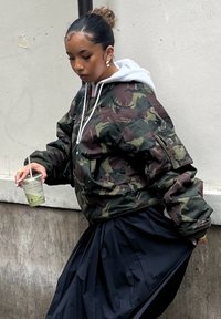 Femme aux cheveux attachés en chignon, portant une veste camouflage et une longue jupe noire, tenant une boisson dans une main en marchant le long d'un mur.