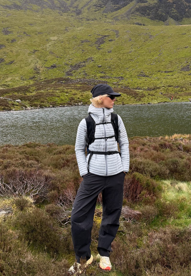 Personne en vêtement d'extérieur et casquette debout au bord d'un lac avec des collines vertes et un terrain rocheux en arrière-plan.