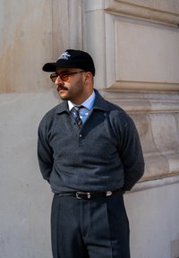 Pullover grigio in lana con design a colletto, cappello nero e occhiali da sole marroni; abbinato con pantaloni scuri e una cravatta a fantasia, in piedi contro un muro.