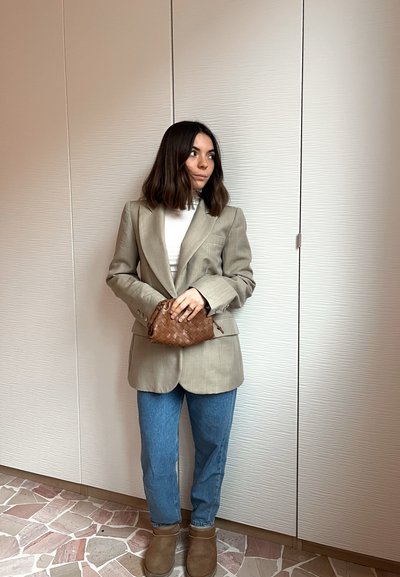 Mujer con blazer beige y suéter de cuello alto blanco sostiene un bolso marrón tejido, viste jeans azules y botas marrones, de pie frente a una pared texturizada.