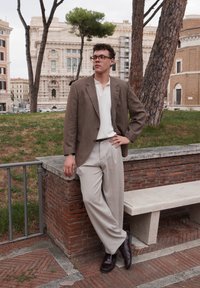 Blazer castanho claro sobre uma t-shirt branca, combinado com calças largas bege e sapatos de couro escuros. De pé contra um fundo de tijolos.