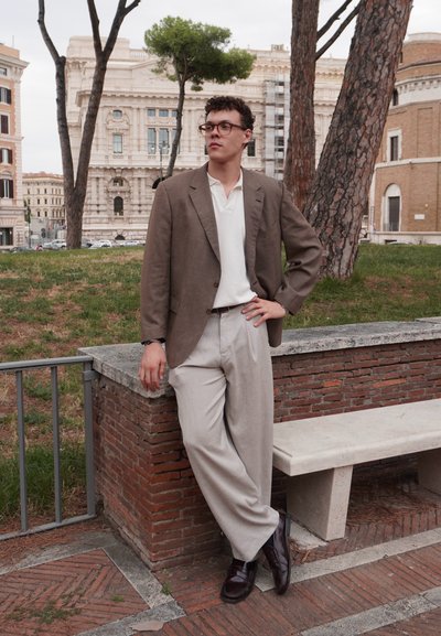 Blazer marrón claro sobre una camiseta blanca, combinado con pantalones beige holgados y zapatos de cuero oscuros. De pie contra un fondo de ladrillo.