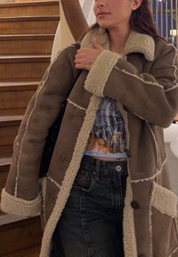 Femme en manteau en peau retournée marron avec garniture crème, debout à l'intérieur près d'un escalier en bois, portant un haut à motifs et un jean foncé.