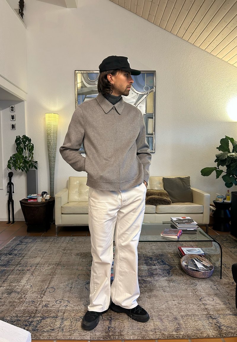 Giacca di lana grigia con colletto, abbinata a pantaloni bianchi e scarpe da ginnastica nere, in piedi in un soggiorno moderno e ben illuminato.
