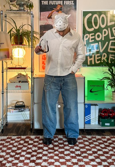 Camisa de rayas blanca de manga larga, jeans holgados azules y zapatos negros. Los accesorios incluyen gafas de sol y una cadena. Fondo con elementos decorativos.