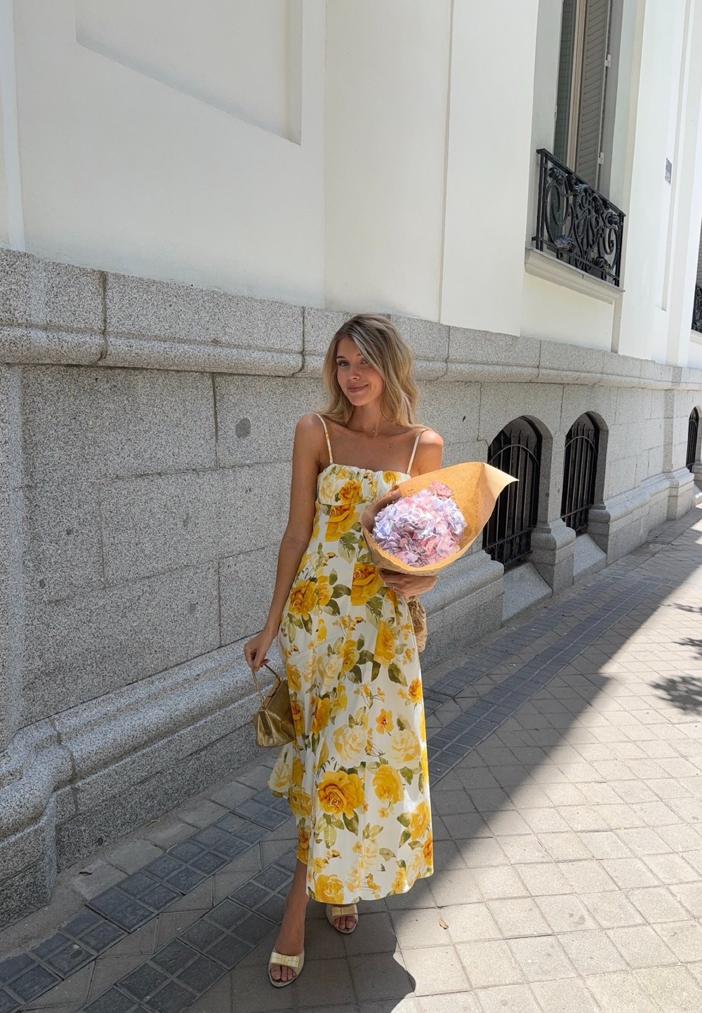 Robe maxi fleurie jaune avec des bretelles spaghetti ; tissu texturé, tenant un bouquet enveloppé dans du papier kraft, debout sur un trottoir pavé.