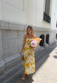 Robe maxi fleurie jaune avec des bretelles spaghetti ; tissu texturé, tenant un bouquet enveloppé dans du papier kraft, debout sur un trottoir pavé.
