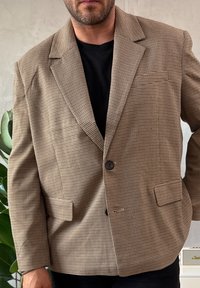 Barna, houndstooth mintás blézer bevágott hajtókával, egysoros gombolással és elején zsebekkel, fekete aláöltözethez párosítva.