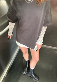 Crna oversized majica s čipkastim dugim rukavima, siva mini suknja i crne gležnjače. Dodaci uključuju zlatni ogrlicu i pametni telefon.