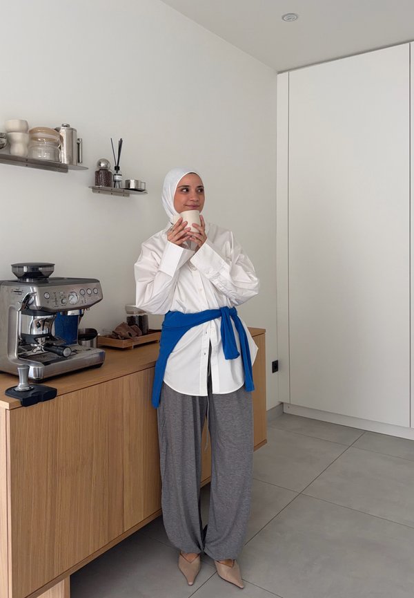 Woman wearing hijab and white shirt with blue sweater tied at waist, holding a cup, standing next to a coffee machine on wooden counter in kitchen.