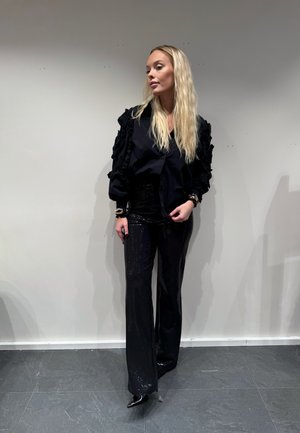 Blusa negra con mangas acampanadas, combinada con pantalones de lentejuelas negros de talle alto. Presenta zapatos de tacón alto y un peinado elegante.