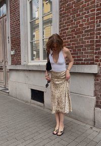Hvit halterneck-top, beige midiskjørt med prikkemønster, svarte sandaler med blomsterdetalj, står på en brosteinsbelagt gate mot en murvegg.