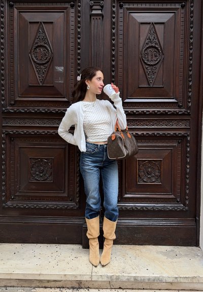 Mujer con cárdigan blanco y blusa de lunares que bebe de una taza, sosteniendo un bolso marrón, de pie frente a una puerta de madera oscura ornamentada.