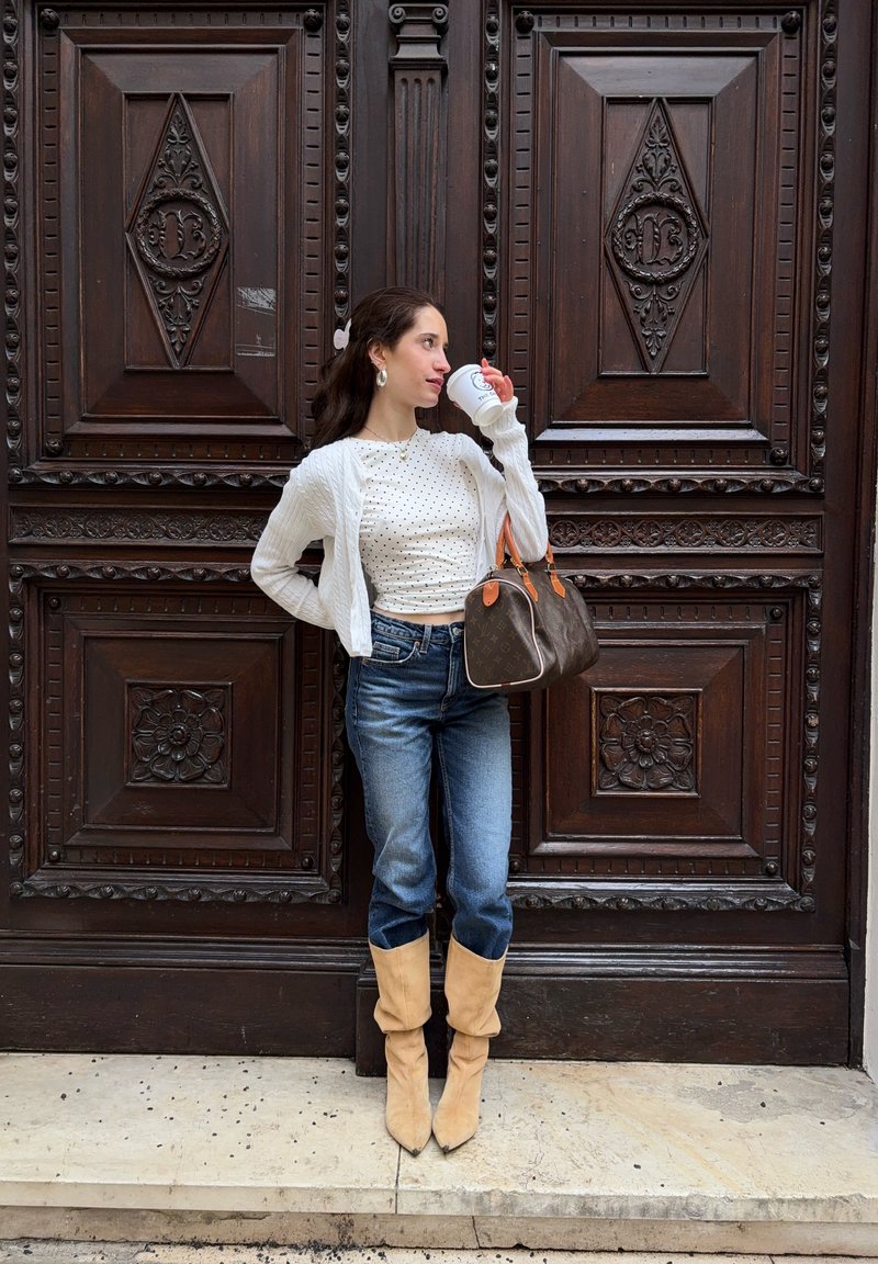 Femme en cardigan blanc et haut à pois buvant dans une tasse, tenant un sac à main marron, debout devant une porte en bois sombre ornée.