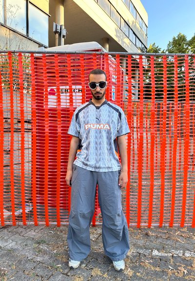 Pantalones grises de corte amplio, camisa de manga corta azul con estampado y logotipo de "PUMA", y gafas de sol blancas contra un fondo de malla naranja.