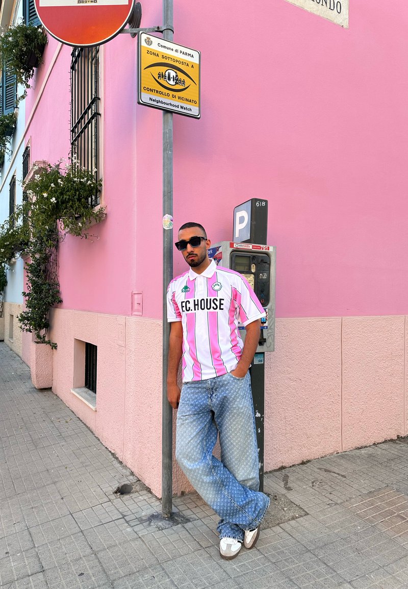 Camisa polo riscada em rosa e branco com logotipo, combinada com calças de ganga azuis claras com padrão. A pessoa está de pé perto de um parquímetro cinza e de uma parede rosa.