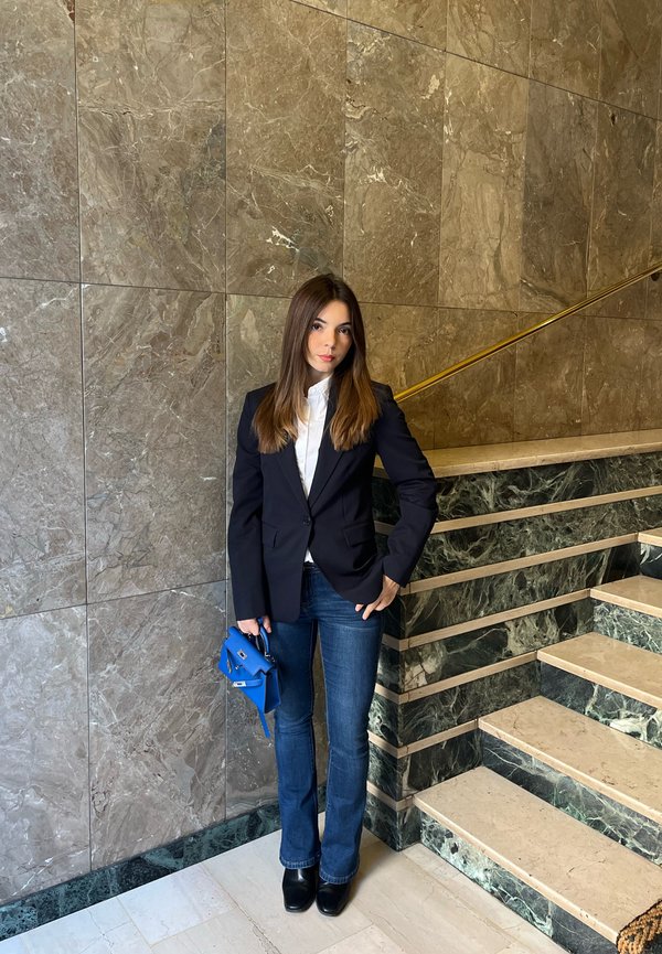 Mörkblå blazer över en vit skjorta, blå handväska och utsvängda jeans. Miljön har marmorväggar och en grön marmortrappa.