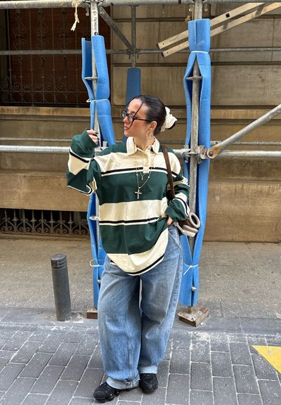 Camisa de rayas verde y crema de manga larga con cuello, combinada con vaqueros azules sueltos y zapatillas negras, de pie frente a un andamiaje.