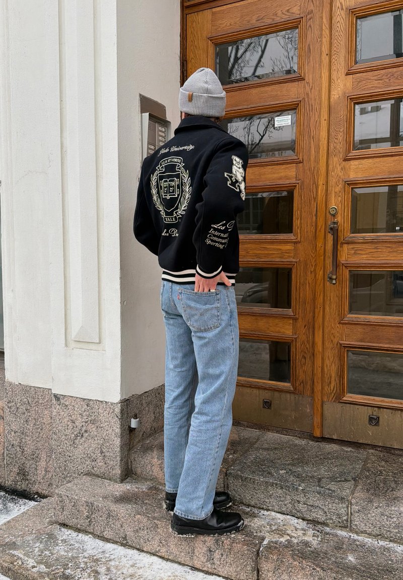 Personne portant un bonnet gris, une veste varsity noire, un jean bleu et des bottes noires, debout devant une porte en bois avec des panneaux en verre.