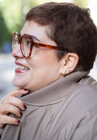 Άτομο με κοντά καστανά μαλλιά που φοράει γυαλιά με σκελετό από χελώνια και πολλά σκουλαρίκια, χαμογελάει και κρατάει το γιακά ενός μπεζ μπουφάν σε εξωτερικό χώρο.