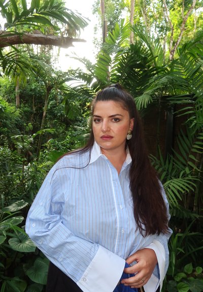 Mujer de cabello largo y oscuro y pendientes dorados, vestida con una camisa azul a rayas, de pie en un frondoso jardín tropical verde.