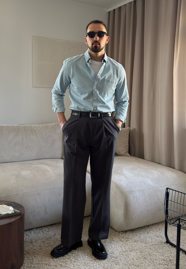 Άνδρας που φοράει γυαλιά ηλίου, ανοιχτό μπλε πουκάμισο με κουμπιά, μαύρη ζώνη, σκούρο παντελόνι και μαύρα παπούτσια, στέκεται σε ένα μοντέρνο σαλόνι.