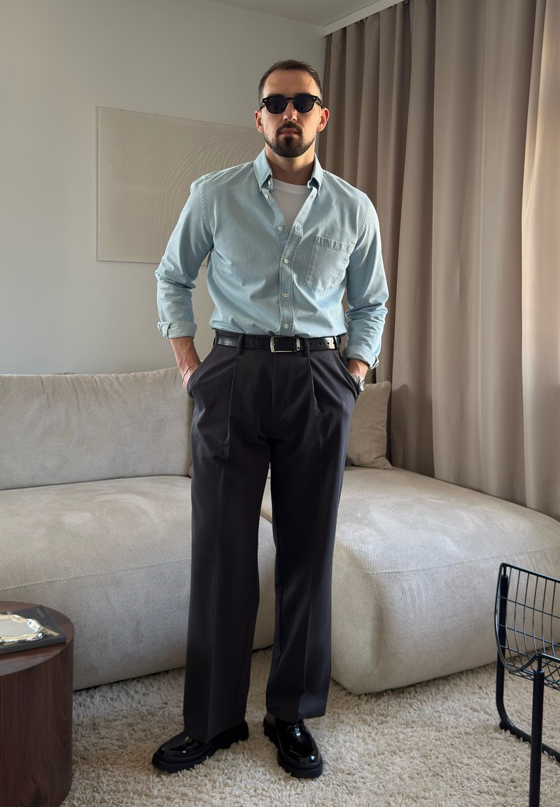 Homme portant des lunettes de soleil, une chemise bleu clair boutonnée, une ceinture noire, un pantalon foncé et des chaussures noires, debout dans un salon moderne.