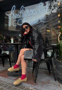 Μαύρο δερμάτινο μπουφάν με φούντες, κόκκινο πουλόβερ, μαύρα γυαλιά ηλίου, μαύρη τσάντα, μπεζ παπούτσια και κόκκινες κάλτσες, καθισμένος σε ένα τραπέζι καφέ.