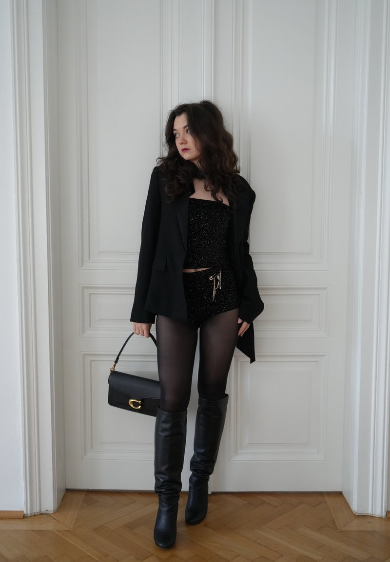Haut et shorts noirs à sequins superposés sous un blazer noir. Bottes noires montantes jusqu'aux genoux et sac à main noir avec garnitures dorées. Sol en bois clair.
