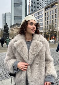 Faux fur-frakke i lys grå med en bred krave. Har en blød tekstur, oversized pasform og detaljeret syning. Bæres med en grå sweater og hat.